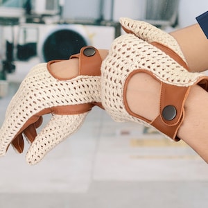 Peut inclure: Une paire de gants de conduite crème avec un motif crocheté. Les gants présentent des accents en cuir marron au poignet et autour des trous pour les doigts, fixés par une fermeture à bouton foncé. Gants de conduite.