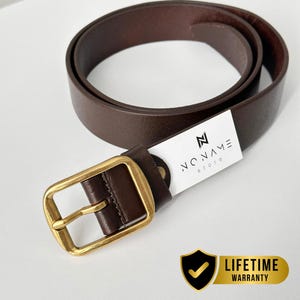 May include: A dark brown leather belt featuring a rectangular brass buckle. A white tag attached to the belt displays the text "N NONAME store". A gold-colored shield icon with a checkmark and the text "LIFETIME WARRANTY" is visible.