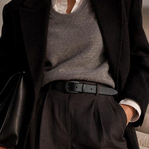 May include: A black blazer worn over a grey crewneck jumper and a white collared shirt. The outfit includes black trousers secured with a black leather belt featuring a matching black buckle, and a black leather handbag held at the side.