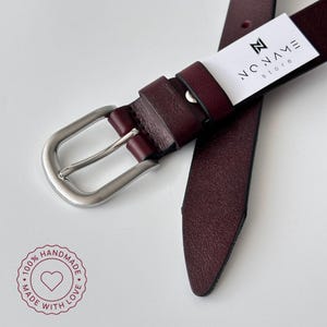 May include: A deep burgundy leather belt featuring a silver-toned metal buckle and a white rectangular tag printed with the text NO NAME store. A circular stamp in the corner displays the text 100% HANDMADE MADE WITH LOVE and a heart icon.