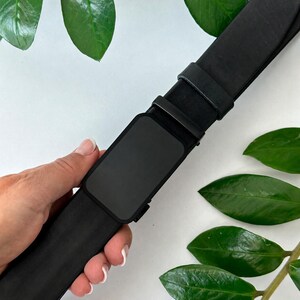 Op de afbeelding: Een effen zwarte riem met een rechthoekige matzwarte gesp en een bijpassende zwarte leren band. De riem heeft een strak en uniform uiterlijk, met een gladde afwerking op zowel de band als de gesp van het hardware-onderdeel.