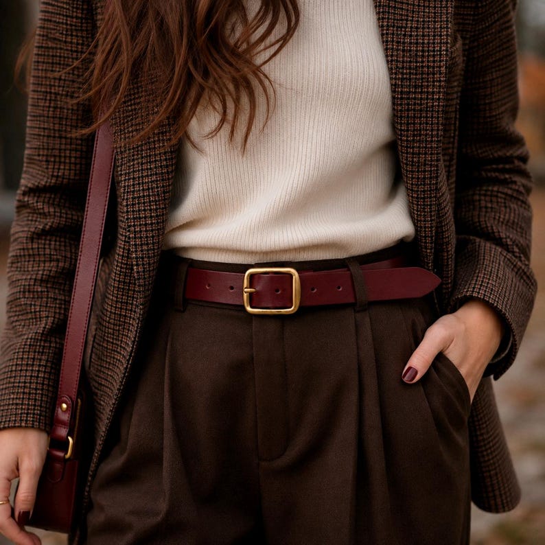 May include: A brown houndstooth blazer worn over a cream ribbed sweater and dark brown trousers. A deep burgundy leather belt with a gold-toned rectangular buckle is fastened at the waist. A matching burgundy leather shoulder bag strap is visible.