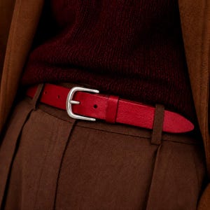 Op de afbeelding: Een rode leren riem met een zilverkleurige metalen gesp, gedragen over een bruine broek en een donkerbordeauxrode trui, met een bruine jas eroverheen. De riem zit door de riemlussen van de bruine broek.