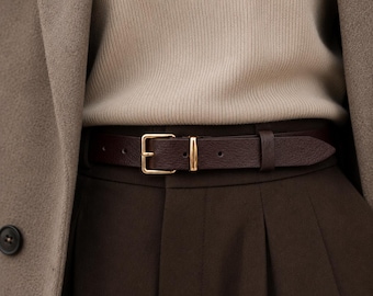 May include: A dark brown leather belt with a gold-toned metal buckle and a matching gold-toned metal keeper loop. The belt is worn over dark brown pleated trousers, paired with a beige ribbed sweater and a light brown wool coat layer.