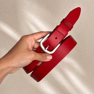 Può includere: Una cintura rosso vivace con finitura liscia e fibbia in metallo color argento opaco. La cintura include un passante in pelle rossa abbinato e fori preforati per la regolazione, caratterizzata da un design pulito, punta arrotondata e cuciture.