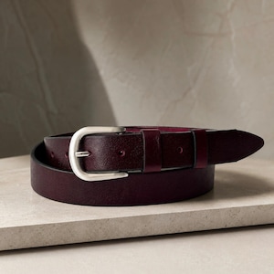 May include: A deep burgundy leather belt featuring a smooth texture and a polished silver-toned metal buckle. The belt is coiled into a circular shape, showcasing the dark red strap, a single silver prong, and a matching leather keeper loop.