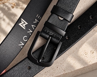 May include: A black leather belt featuring a matte black metal buckle and two matching keepers. The strap has a textured surface and displays the white text NO NAME with a stylized N logo printed clearly on the leather near the buckle assembly.