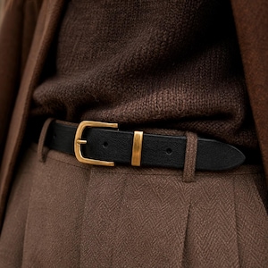 May include: A black belt featuring a gold-toned metal buckle and a matching gold-toned metal keeper loop. The belt is worn over brown trousers and a brown knitted jumper, with a brown jacket layered over the top of the outfit.