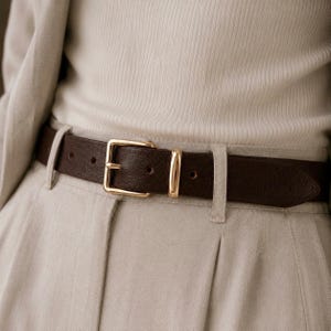 May include: A dark brown leather belt with a polished gold-tone metal buckle and a matching rectangular keeper loop. The belt is threaded through the belt loops of light beige trousers, worn over a ribbed cream-colored top and a light beige blazer.