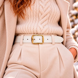 May include: A white leather belt with a rectangular brass-toned buckle, worn over cream-colored pleated trousers and a matching cable-knit sweater, layered beneath a long, light beige coat.