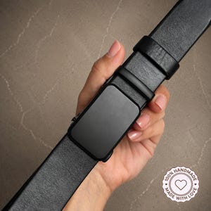 May include: A black leather belt featuring a smooth, rectangular matte black buckle and a matching leather keeper loop. The belt strap has a visible grain texture and includes a circular stamp that reads 100% HANDMADE MADE WITH LOVE.