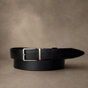 Peut inclure: Une ceinture noire avec une finition lisse et une boucle rectangulaire en métal de couleur bronze à canon. La sangle est enroulée, présentant un passant en cuir noir assorti près de la boucle, avec une extrémité pointue vers la droite.