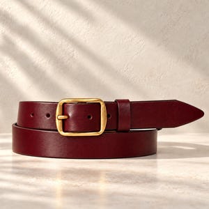 May include: A deep burgundy leather belt featuring a smooth finish and a rectangular brushed gold-tone metal buckle. The belt includes a matching leather keeper loop and a pointed tip, showcasing a clean and simple design with visible holes.