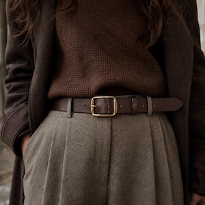 May include: A dark brown leather belt with a rectangular brass buckle worn over pleated taupe trousers, a dark brown knit sweater, and a dark brown coat. The belt features a single prong and a matching leather keeper loop for a secure fit.