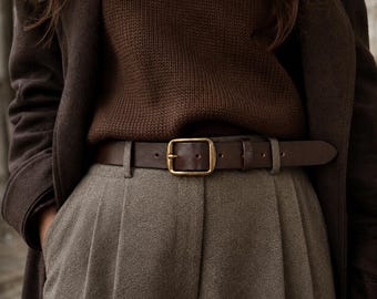 May include: A dark brown leather belt with a rectangular brass buckle, worn over pleated taupe trousers and a dark brown knit sweater, layered beneath a dark brown coat. The belt features a simple pin closure and a single leather loop.