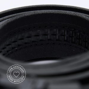 May include: A black leather belt coiled in a circular shape, featuring a textured inner surface with a repeating square pattern and visible stitching along the edges. A circular white logo with a heart icon and the text 100% HANDMADE MADE WITH LOVE.