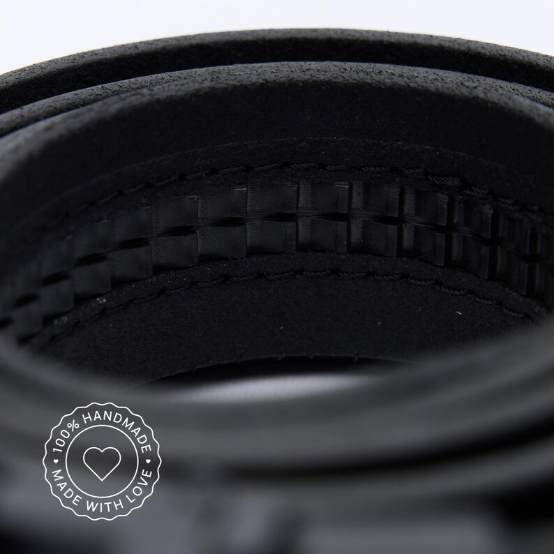 May include: A black leather belt coiled in a circular shape, featuring a textured inner surface with a repeating square pattern and visible stitching along the edges. A circular white logo with a heart icon and the text 100% HANDMADE MADE WITH LOVE.