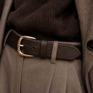 May include: A dark brown leather belt featuring a polished gold-tone metal buckle. The belt is threaded through the belt loops of light brown trousers, worn over a dark brown knit sweater and a textured brown herringbone blazer jacket.