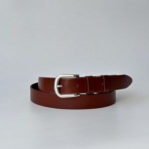 May include: A smooth, dark brown leather belt featuring a polished silver-tone metal buckle. The belt is coiled in a circular shape, showcasing a single prong, a rectangular frame, and two matching leather keepers along the strap.