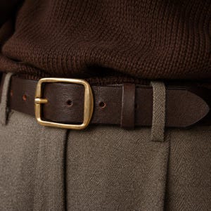 May include: A dark brown leather belt featuring a rectangular brass buckle. The belt is threaded through the belt loops of light brown herringbone trousers, positioned below a dark brown knitted sweater.