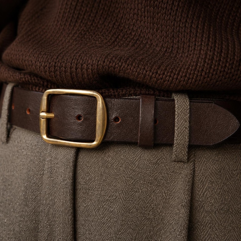 May include: A dark brown leather belt featuring a rectangular brass buckle. The belt is threaded through the belt loops of light brown herringbone trousers, positioned below a dark brown knitted sweater.
