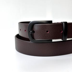 May include: A dark brown belt featuring a matte black metal buckle and a matching black belt loop. The belt strap has a smooth, textured surface with visible circular adjustment holes that reveal a deep magenta or dark pink interior lining.