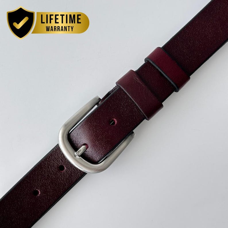 May include: A deep burgundy leather belt featuring a smooth texture, a silver-toned metal buckle, and two matching leather keepers. A gold shield-shaped emblem in the corner displays a checkmark and the text LIFETIME WARRANTY.