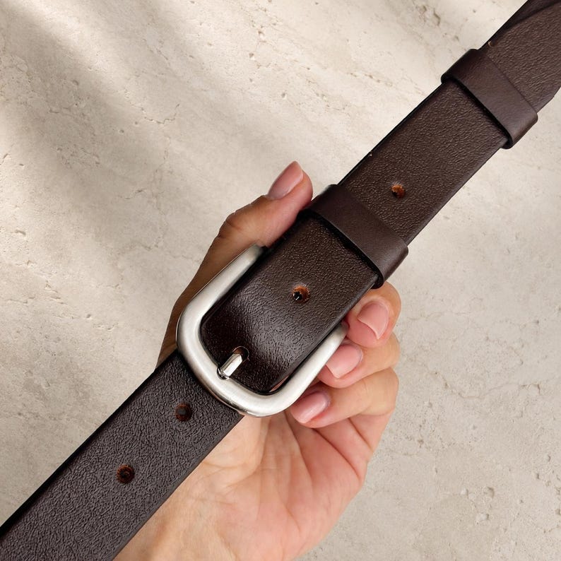 May include: A dark brown leather belt featuring a smooth, rounded silver-tone metal buckle. The belt strap has a textured surface with evenly spaced circular holes and a matching leather keeper loop to secure the end of the strap in place.