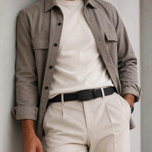 May include: A taupe long-sleeved button-down shirt worn over a cream-colored crew neck t-shirt, paired with light beige trousers and a black belt featuring a rectangular buckle. The shirt has two chest pockets and dark buttons down the front.