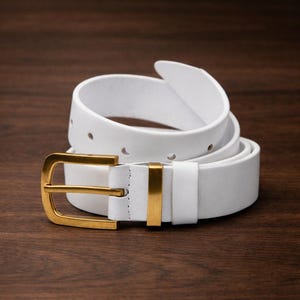 Puede incluir: Un cinturón de cuero blanco con una hebilla de metal dorado pulido y una trabilla de metal dorado a juego. El cinturón está enrollado, mostrando un acabado liso, una serie de agujeros perforados para el ajuste y una trabilla de cuero blanco.