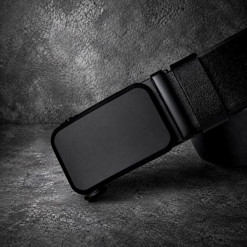 May include: A black belt featuring a rectangular, matte black buckle with a smooth, solid surface. The strap has a textured finish and is attached to the buckle mechanism, which includes a small side release lever for adjustment.