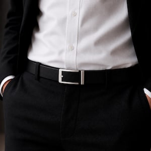 Puede incluir: Un cinturón de cuero negro con una hebilla rectangular de metal plateado. El cinturón se lleva alrededor de la cintura sobre un pantalón negro y una camisa blanca abotonada, parcialmente cubierta por una chaqueta negra.