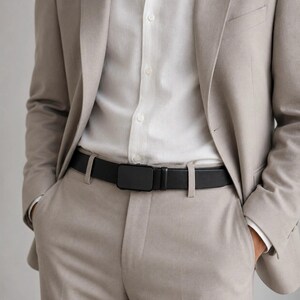 May include: A black leather belt with a rectangular matte black buckle worn over a white button-down shirt and a light beige blazer and matching trousers. The belt features a smooth finish and a simple, clean design with a single belt loop.