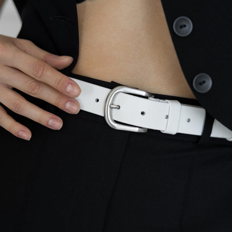 May include: A white belt with a smooth finish and a matte silver-toned metal buckle. The belt features a standard prong closure and a matching white leather-like keeper loop, worn through the belt loops of black trousers under a black blazer.