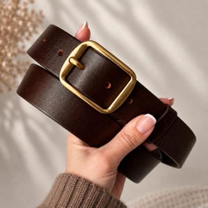 May include: A dark brown leather belt featuring a smooth, matte finish and a rectangular gold-toned metal buckle. The belt strap has visible adjustment holes and a matching leather keeper loop, showcasing a clean and simple design.