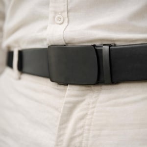 May include: Close-up of a black belt with a matte finish and a rectangular buckle. The belt is worn over a light-colored button-down shirt and pants. The buckle is a simple, modern design.