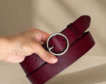 May include: A deep burgundy belt featuring a smooth, matte finish and a circular silver-toned metal buckle. The belt strap is constructed from a solid material with visible stitching along the edges and pre-punched holes for an adjustable fit.