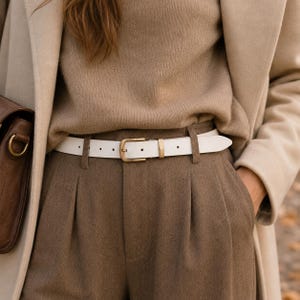 May include: A white belt with a gold-toned metal buckle and a matching gold-toned keeper loop, worn through the belt loops of brown trousers. The belt is positioned over a tan jumper and beneath a beige coat, with a brown bag visible on the side.