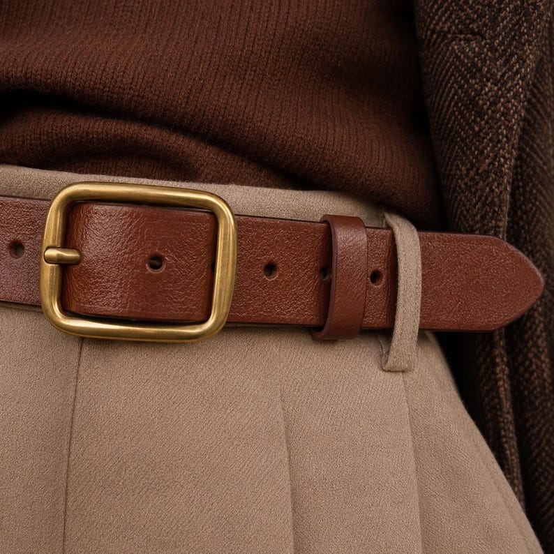 May include: A brown leather belt featuring a rectangular brass buckle with a rounded edge. The belt is worn over beige trousers and a brown knit top, with a brown textured blazer visible on the side, showcasing a classic, clean design.
