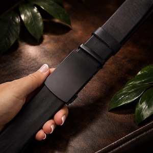 Peut inclure: Une ceinture en cuir noir avec une boucle noire mate. La ceinture est tenue par une main, mettant en valeur la texture lisse du cuir et le design épuré de la boucle. Le fond est une surface en cuir marron foncé.