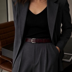 May include: A dark burgundy leather belt with a silver-toned metal buckle worn over high-waisted charcoal gray pleated trousers. The belt is paired with a black ribbed v-neck top and a matching charcoal gray blazer, creating a layered ensemble.