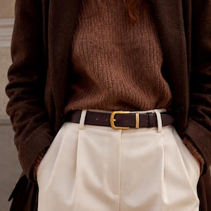 May include: A dark brown leather belt with a matte gold-toned buckle worn with cream-coloured pleated trousers, a brown ribbed knit jumper, and a dark brown coat. The belt features a classic prong closure and a single matching metal keeper loop.