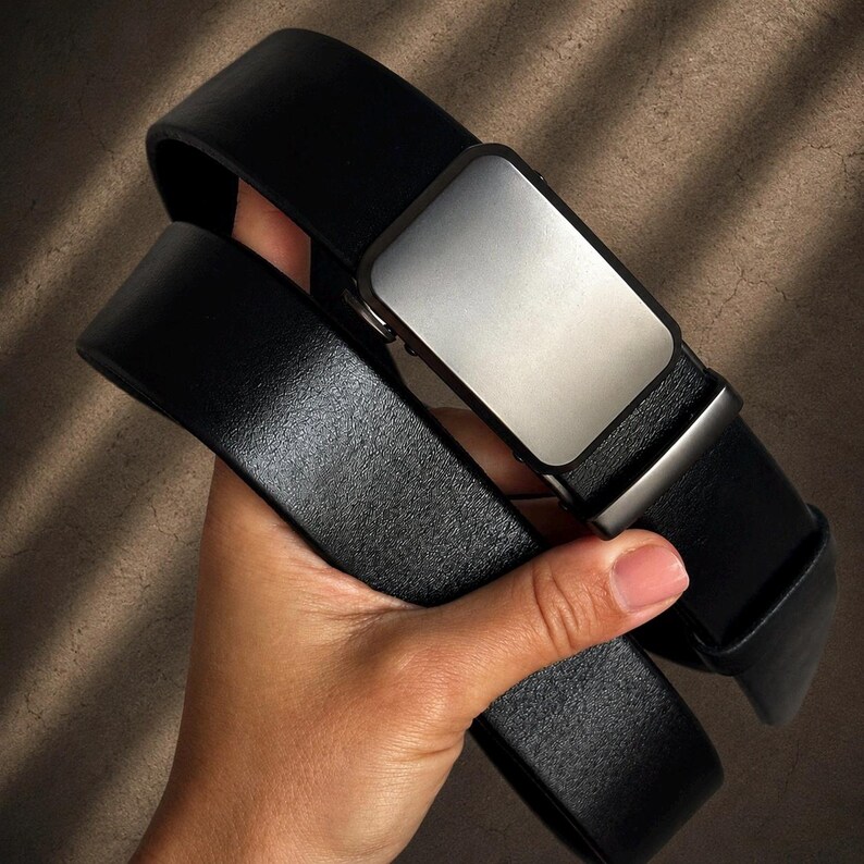 May include: A black belt featuring a smooth, rectangular matte silver buckle. The belt strap is made of a textured black material with a consistent width and a matching black loop near the buckle, showcasing a clean and functional design.