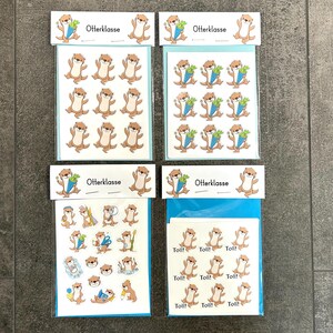 Könnte beinhalten: Vier Stickerpackungen mit Cartoon-Ottern. Einige Otter halten Karotten, andere haben den Text "Toll!" oder sind in verschiedenen Posen. Die Aufkleber sind auf weißen und blauen Trägerkarten verpackt, mit dem Text "Otterklasse" oben.
