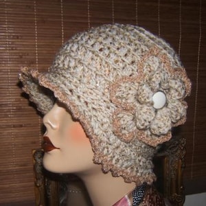 May include: A crocheted hat with a flower design in beige and brown colors. The hat has a scalloped edge and a button accent.