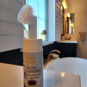 May include: A white bottle of facial cleansing gel with a pump dispenser and a brush attachment. The bottle is labeled "Facial Cleansing Gel with Argan Oil & Saffron" and "For all skin types e 200 ml".