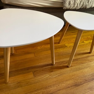 May include: Two white oval-shaped side tables with wooden legs. The larger table is in the foreground and the smaller table is behind it. Both tables are on a wooden floor.