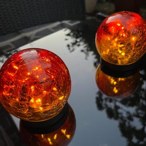 May include: Two illuminated, spherical glass orbs with a crackle effect, displaying a gradient of red to orange hues. The orbs sit on a reflective surface, creating a mirrored effect. The orbs are likely decorative garden lights.