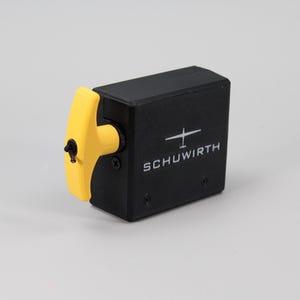 Puede incluir: Un dispositivo mecánico rectangular de color negro con una manija de tracción amarilla brillante en forma de T en el lateral. La parte frontal muestra un gráfico blanco de un planeador sobre el texto SCHUWIRTH en una fuente blanca sin remates.