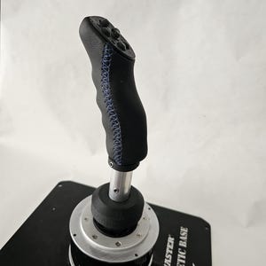 Può includere: Un cambio marcia nero rivestito in pelle con cuciture blu e una base argentata. La base ha la scritta "Thrustmaster" e "Magnetic Base" stampata su di essa.